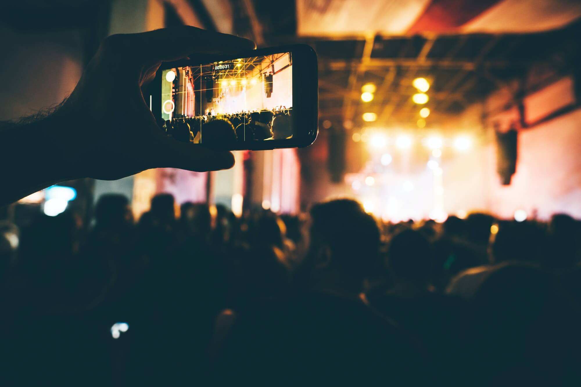 Close up hand with phone at the concert hall. Crowd at the concert and blurred stage lights.